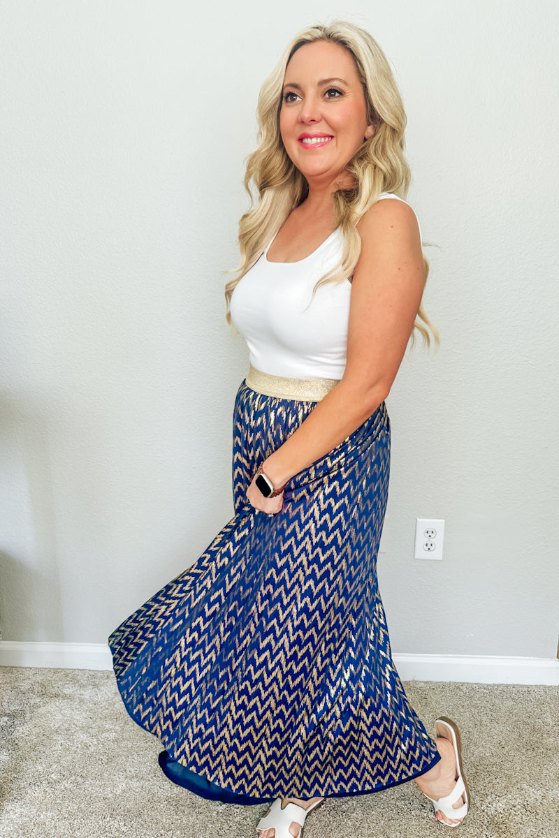 Trendy boutique resort wear featuring a blonde woman twirling in a metallic blue and gold pleated maxi skirt with an elegant gold waistband from aomen's boutique.