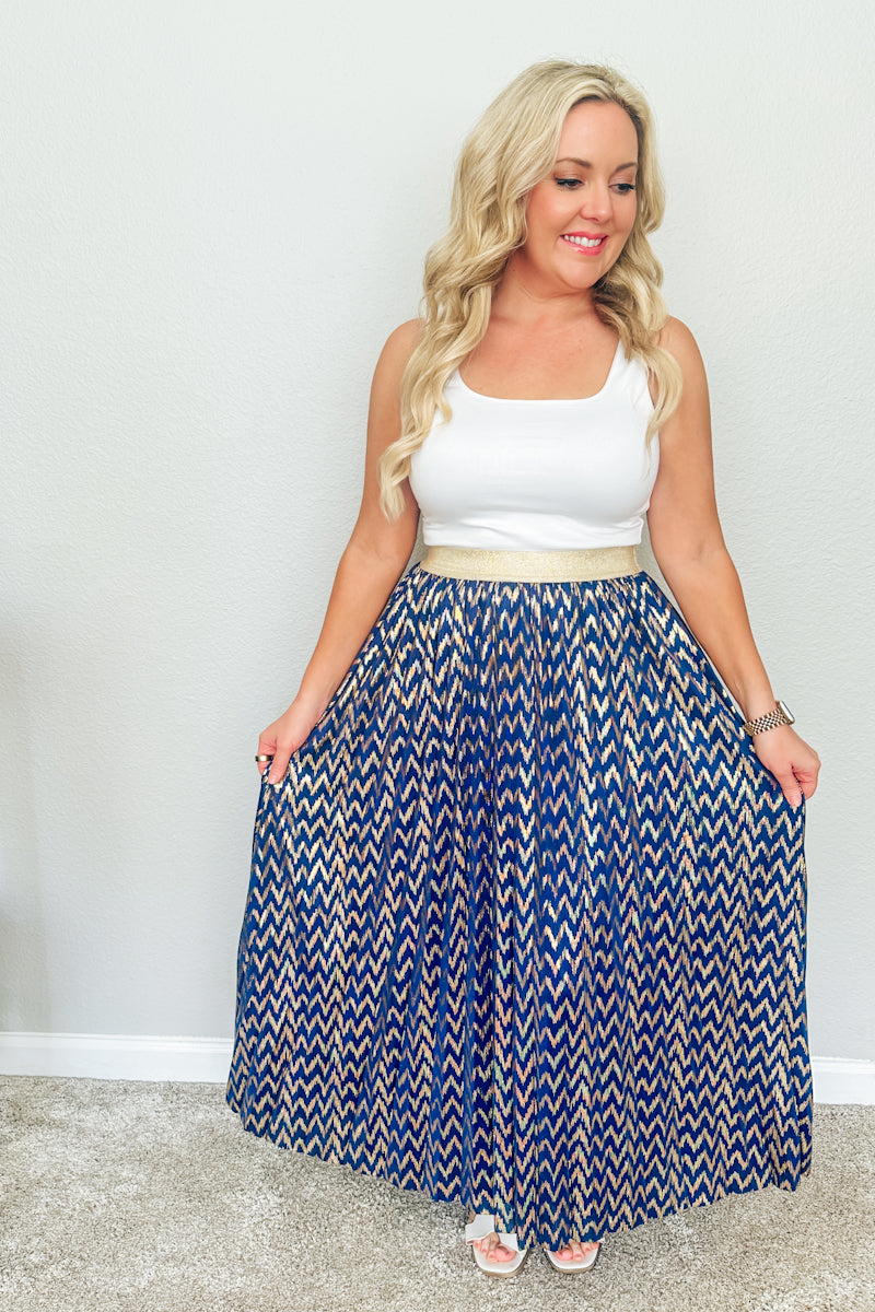 Blonde woman holding out the sides of a flowy blue and gold chevron pleated maxi skirt, demonstrating the elegant movement of this resort wear skirt.