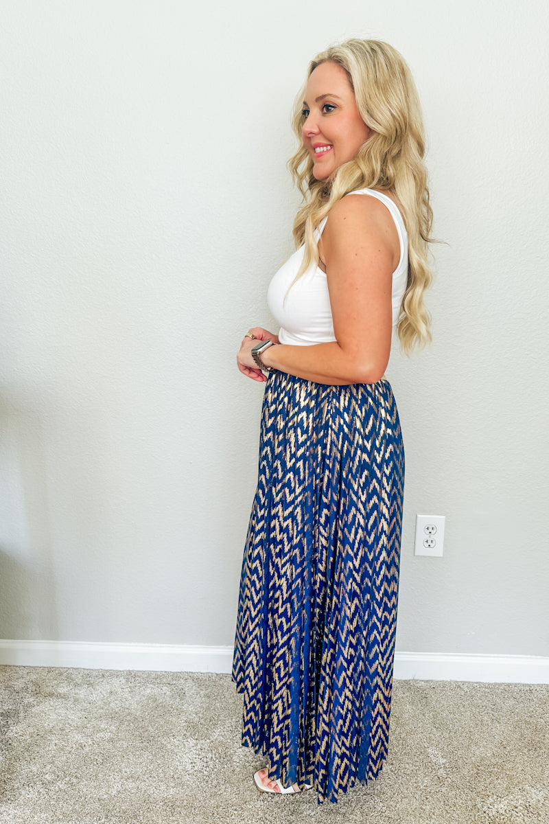 Side profile view of a woman styling a metallic blue and gold pleated maxi skirt with a white bodysuit, showcasing high-quality resort wear fashion.